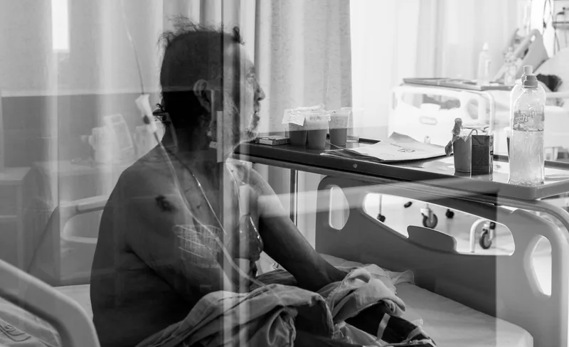 Una persona hospitalizada sentada en su cama, vista a través de una cortina